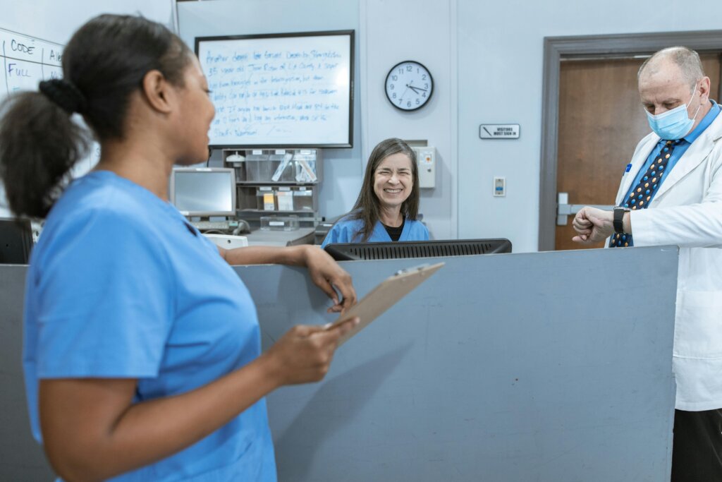 Two nurses in blue scrubs smile at each other over a desk in a healthcare setting while a doctor checks his watch
