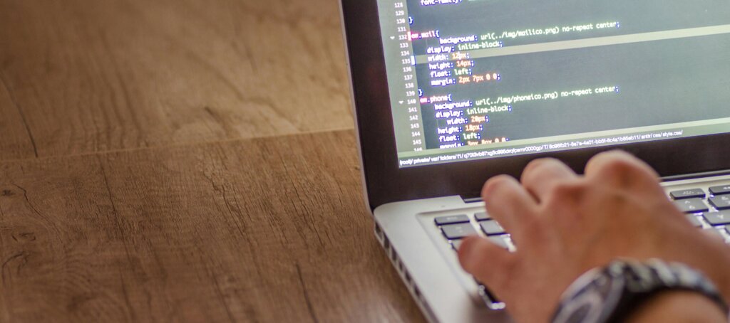 A hand rests on a silver laptop and types computer code which is visible on the laptop screen